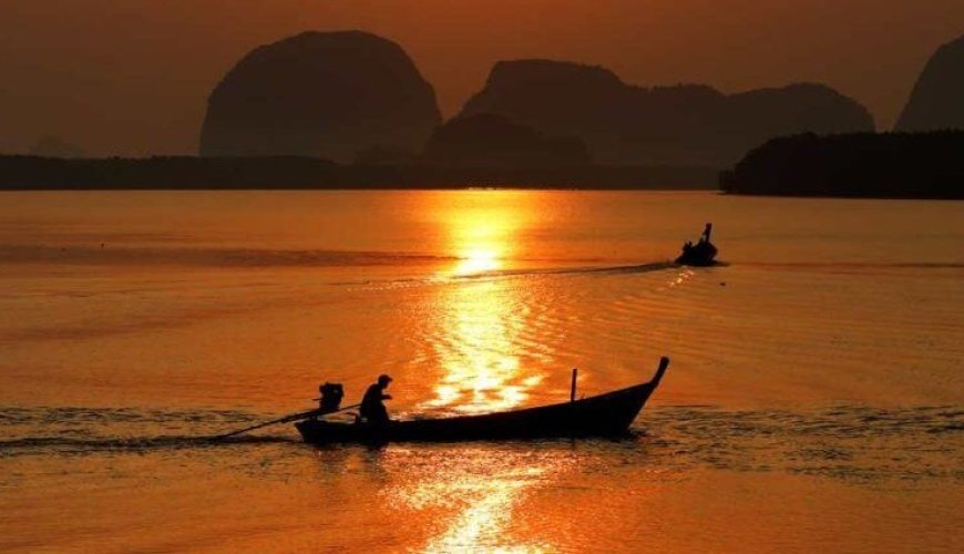A man rows a longtail boat at sunset, with mountains framing the background.